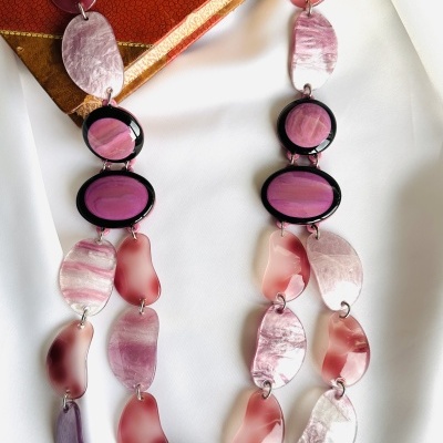 Colar rosa e roxo com peças ovais em padrão marmoreado colocado sobre tecido branco e livro antigo