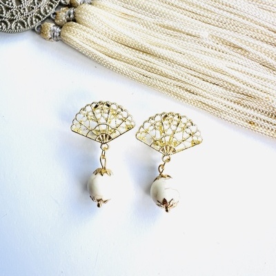 Brincos dourados em forma de leque com pérolas artificiais em fundo branco e franja branca com contas douradas