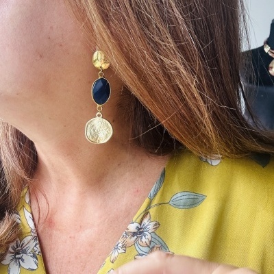 Brinco pendente dourado com pedra preta usado por pessoa com vestido amarelo floral.