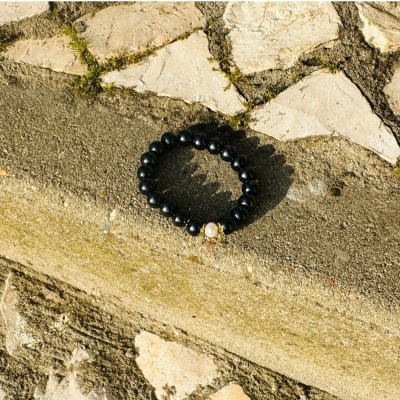 Pulseira elástica de contas negras com uma conta dourada sobre meio-fio de cimento e pedras claras.