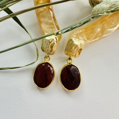 Brincos dourados com pendente oval vermelho escuro sobre fundo branco com ramos secos