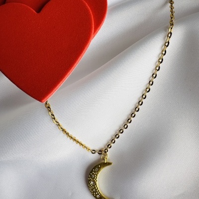 Colar dourado com pendente de lua e pedras brilhantes sobre tecido branco e corações vermelhos