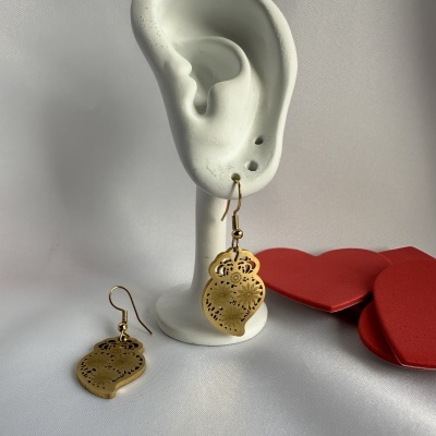 Pendentes dourados em forma de folha com detalhes florais recortados num suporte orelha branca com corações vermelhos ao fundo.