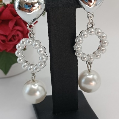 Brincos prateados com pérolas pendurados num suporte preto e rosa vermelha ao fundo