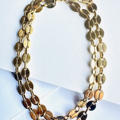 Colar triplo dourado de discos metálicos polidos sobre fundo branco e envelope