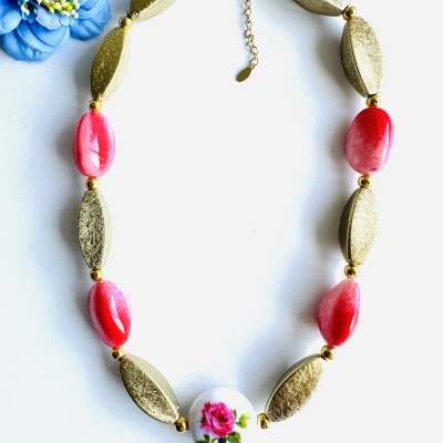 Colar com contas douradas e rosadas e peça central branca com rosa, sobre fundo branco e flor azul