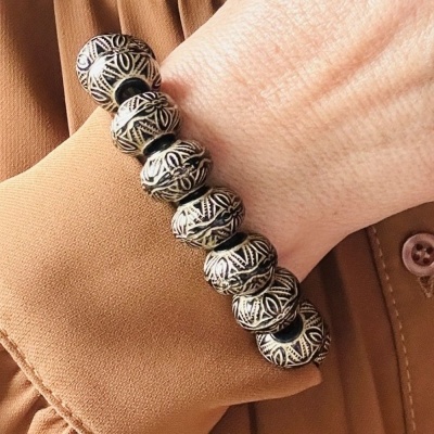 Pulseira de contas metálicas e pretas com padrão geométrico usada no pulso de pessoa com camisa castanha clara
