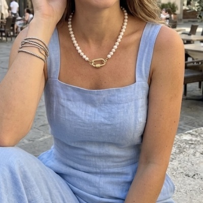 Pessoa com vestido azul e colar de pérolas com fecho dourado, pulseiras prateadas, sentada em ambiente exterior