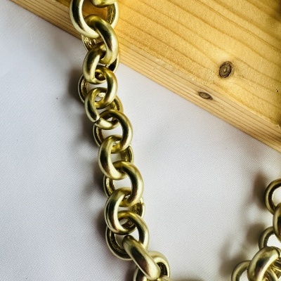 Colar cadeia grossa dourada com elos ovais e redondos sobre superfície branca e madeira clara.