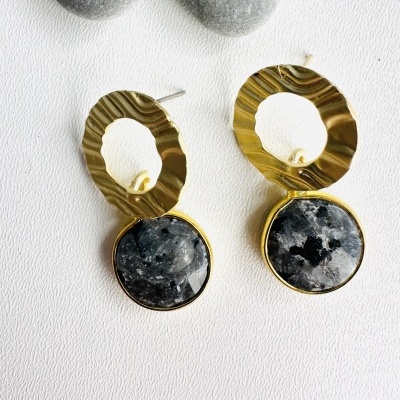 Brincos dourados com pedras cinzentas sobre fundo branco e pedras lisas ao fundo.