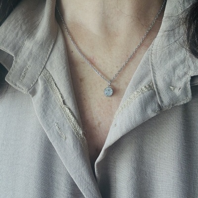 Colar prateado com pendente redondo numa pessoa com camisa cinza.