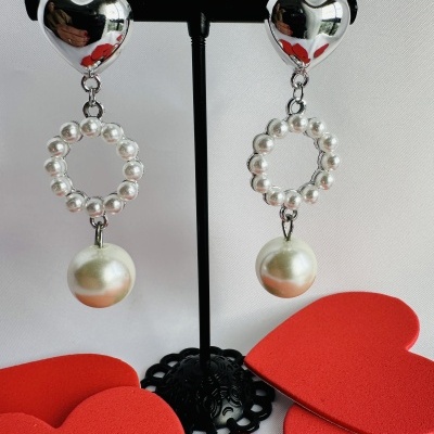 Brincos com formato de coração prateado, círculo de pérolas e pérola pendurada em suporte preto com corações vermelhos ao redor