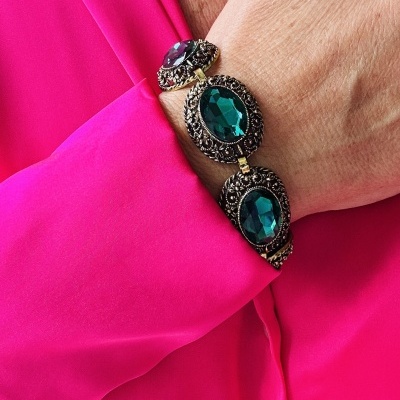 Pulseira com pedras verdes e roxas em armação metálica trabalhada no pulso com camisa rosa