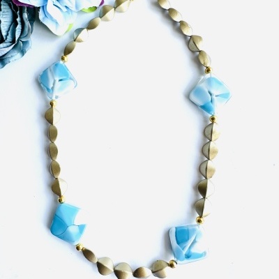 Colar metálico dourado com pedras azuis claras sobre fundo branco e flores azuis