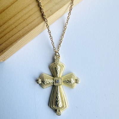 Colar com pendente em forma de cruz dourada detalhada sobre fundo branco e madeira.