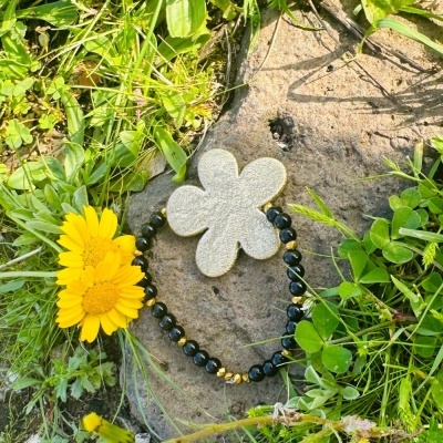 Pulseira com contas pretas e douradas e pendente de flor cinzenta sobre pedra e relva com flor amarela