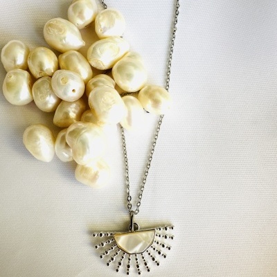 Colar com corrente metálica e pendente semi-circular com pérolas e esferas metálicas junto a pérolas marfim irregulares sobre tecido branco.