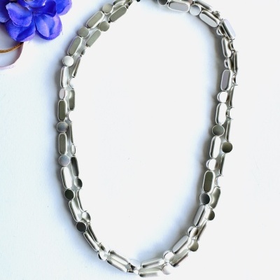 Colar de metal prateado com elos alongados em fundo branco e flor azul ao lado