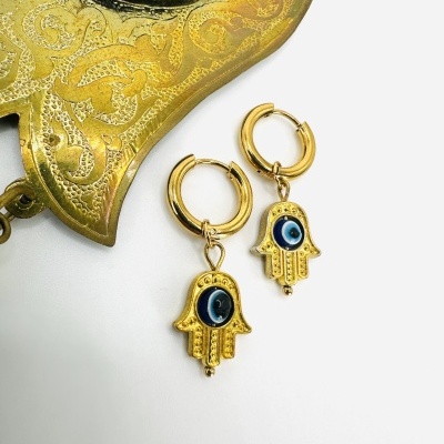 Brincos dourados com pendente em forma de mão Hamsá e olho azul sobre superfície branca e objeto dourado decorativo