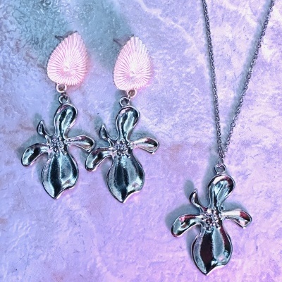 Conjunto de brincos e colar com pendentes prateados em forma de flor sobre fundo lilás