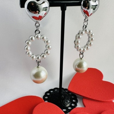 Brincos pendentes com coração de metal prateado e pérolas brancas em suporte preto com corações vermelhos à volta.