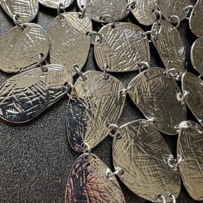 Colar de placas metálicas prateadas texturadas ligado por argolas sobre fundo preto.