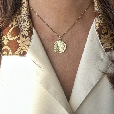 Colar dourado com medalha redonda em pescoço com blusa branca e lenço castanho.
