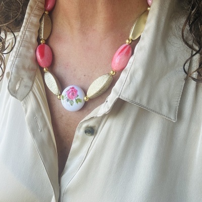 Colar artesanal com contas ovais rosa e douradas e peça central branca com flor rosa, em blusa bege clara