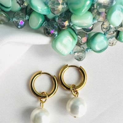 Brincos dourados com pérolas penduradas sobre tecido branco e pedras verdes ao fundo