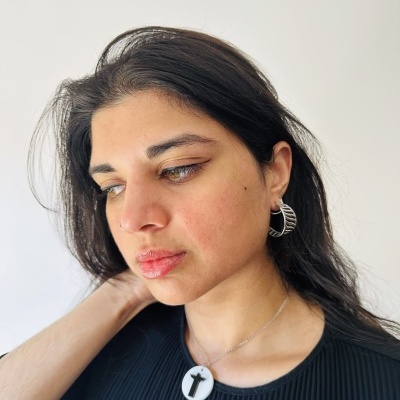 Mulher com colar de pendente redondo branco com cruz preta e brincos prateados em anel, vestindo camisa preta, fundo branco