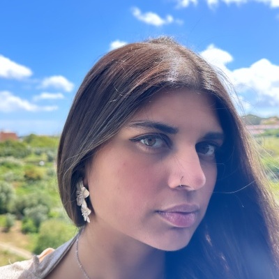 Mulher com brinco de concha e colar prateado ao ar livre, céu azul e vegetação ao fundo