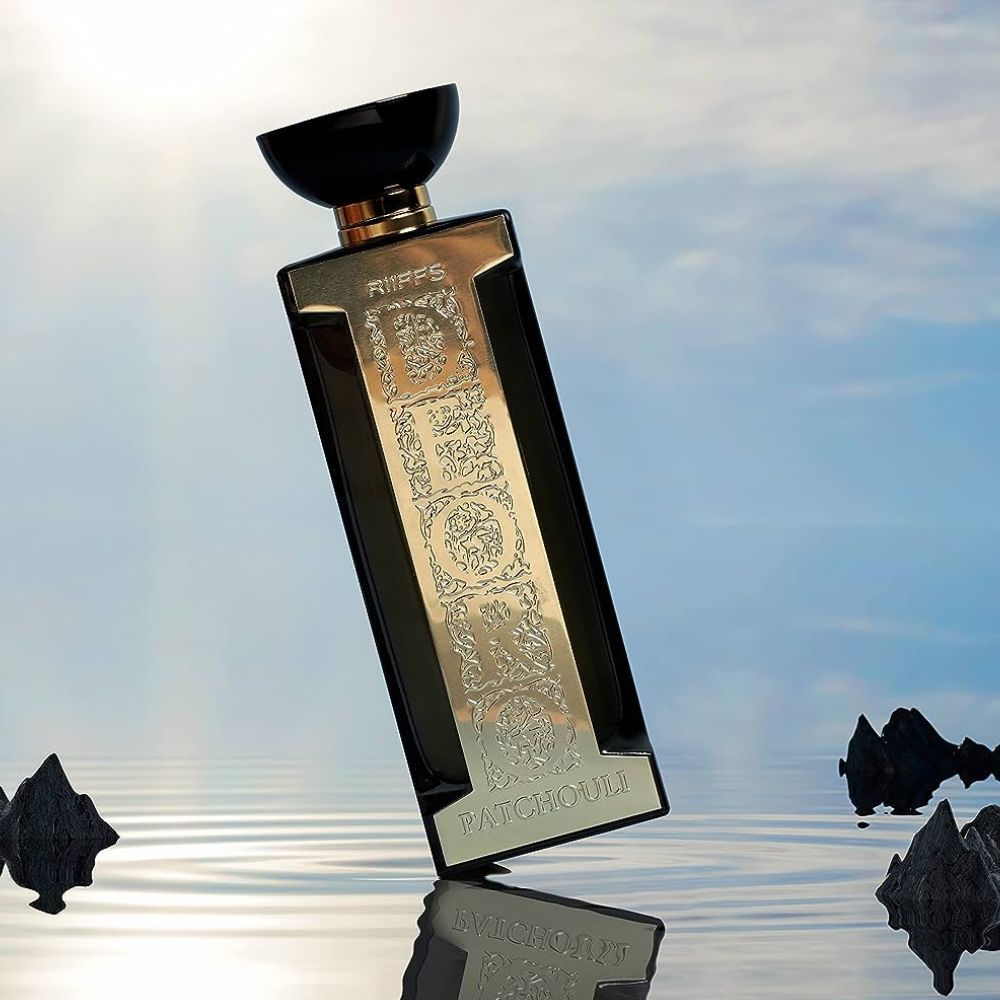 Frasco de perfume preto e dourado sobre superfície reflexiva com fundo de céu azul e nuvens