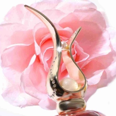 Frasco de perfume com tampa dourada decorada e flor rosa ao fundo.