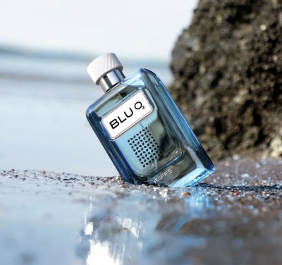 Frasco de perfume azul sobre areia molhada com rocha ao fundo