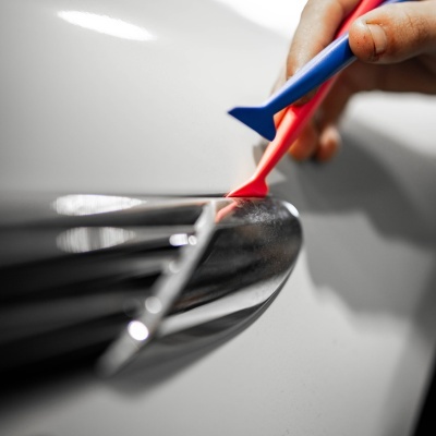 Mão a aplicar adesivo preto brilhante num carro branco com ferramentas plásticas