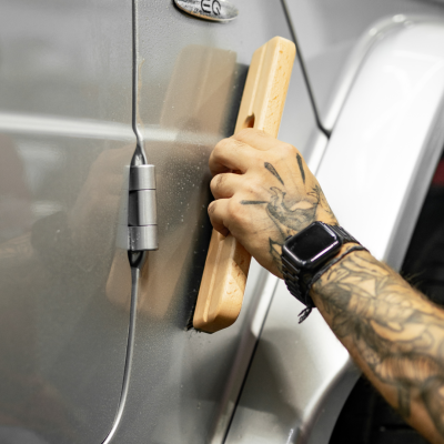 Mão com tatuagens segurando esponja de madeira numa porta de carro cinza