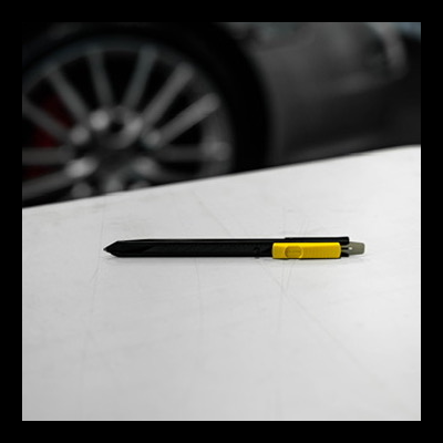 Caneta preta com detalhe amarelo sobre mesa branca com roda de carro ao fundo