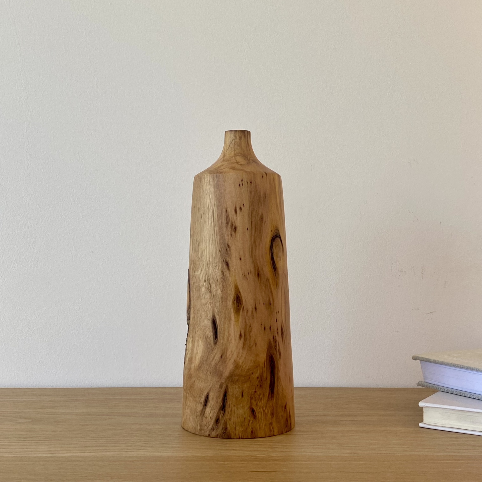 JARRA EM MADEIRA DE CIPRESTE Vaso alto de madeira natural com textura visível, sobre mesa de madeira clara ao lado de livros brancos