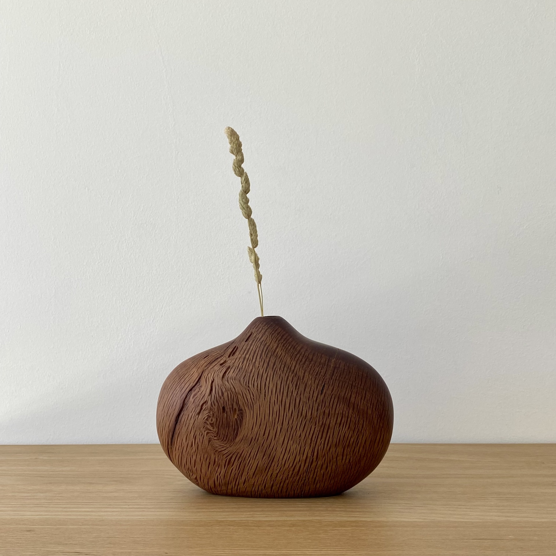 JARRA EM MADEIRA DE AZINHO Vaso de madeira castanho com ramo seco, sobre mesa de madeira clara, parede branca ao fundo