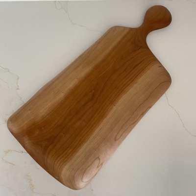 Tábua de cortar em madeira clara com cabo, sobre bancada branca marmoreada