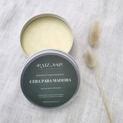 Frasco metálico de cera para madeira aberto com tampa preta e texto branco sobre tecido cinza claro.