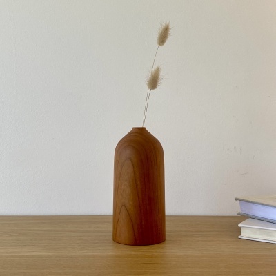 Vaso alto em madeira com duas hastes secas, sobre mesa de madeira e fundo branco.