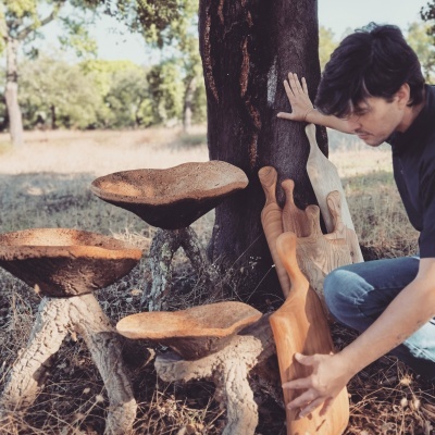 Três bancos ou mesas baixas de madeira natural com suportes texturizados, homem segurando tábuas esculpidas ao lado de uma árvore ao ar livre