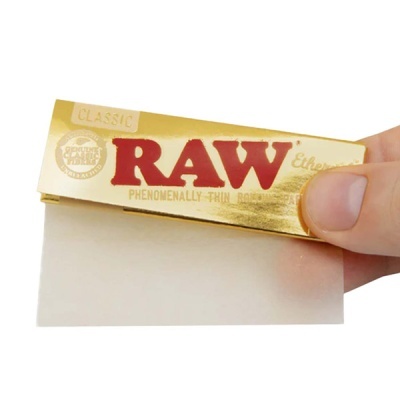 Folha de papel RAW Classic dourada com texto em vermelho e dourado, segurada por mão humana