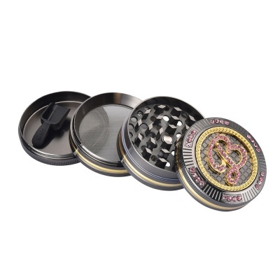 Triturador de metal cinza escuro com detalhes em dourado e pedras rosa, e raspador preto
