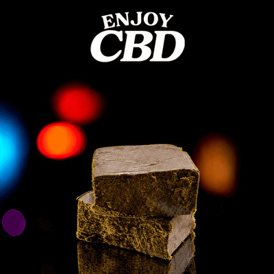 Dois pedaços quadrados castanhos com textura em fundo preto com luzes coloridas e texto ENJOY CBD