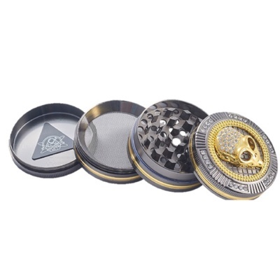 Triturador metálico prateado e dourado com caveira decorativa no topo e espátula triangular preta