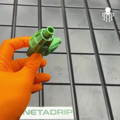 Componente verde plástico segurado por mão com luva laranja sobre ladrilhos cinza, texto NETADRIP ao fundo