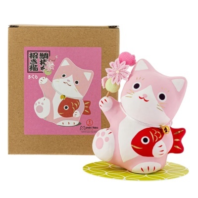 Figura de gato da sorte cor-de-rosa e branco com peixe vermelho e flores, ao lado de caixa castanha com etiqueta rosa.
