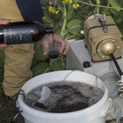 Homem despejando líquido preto em balde branco com líquido escuro e máquina mecânica dourada em ambiente com plantas
